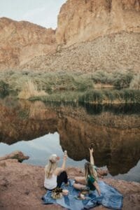 Two women enjoying a picnic by a serene lake with rocky canyon views.